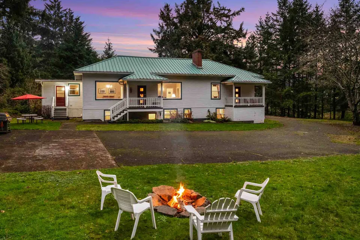 The image showcases the historic Manzanita Schoolhouse nestled among tall trees, with a glowing fire pit surrounded by outdoor seating in the spacious yard, inviting guests to gather and relax. The house's classic architecture is warmly lit, and the natural setting exudes both comfort and privacy, perfect for group getaways.