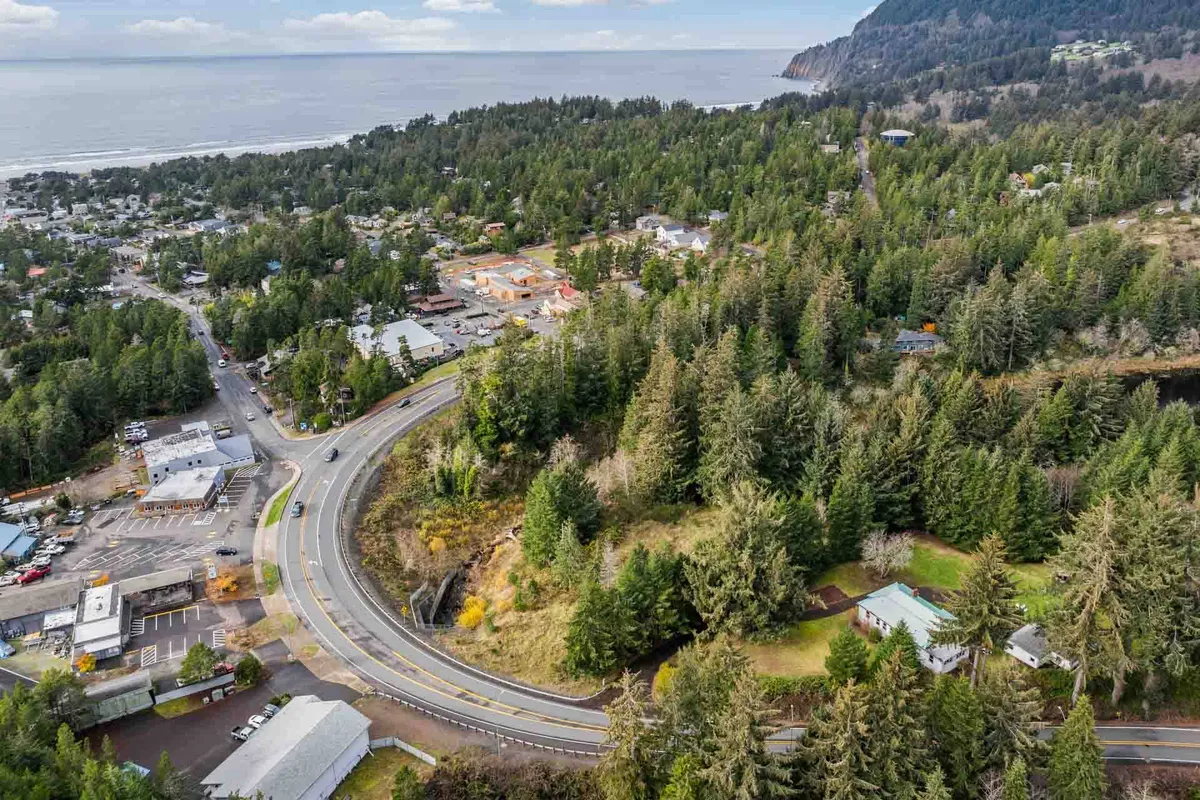 This image captures a panoramic view of the Oregon Coast near Manzanita, featuring a winding coastal road bordered by dense forest, a glimpse of the town, and the expansive ocean beyond. The tranquil setting highlights the proximity to the beach and the region’s natural beauty, with hints of sunset light over the water.