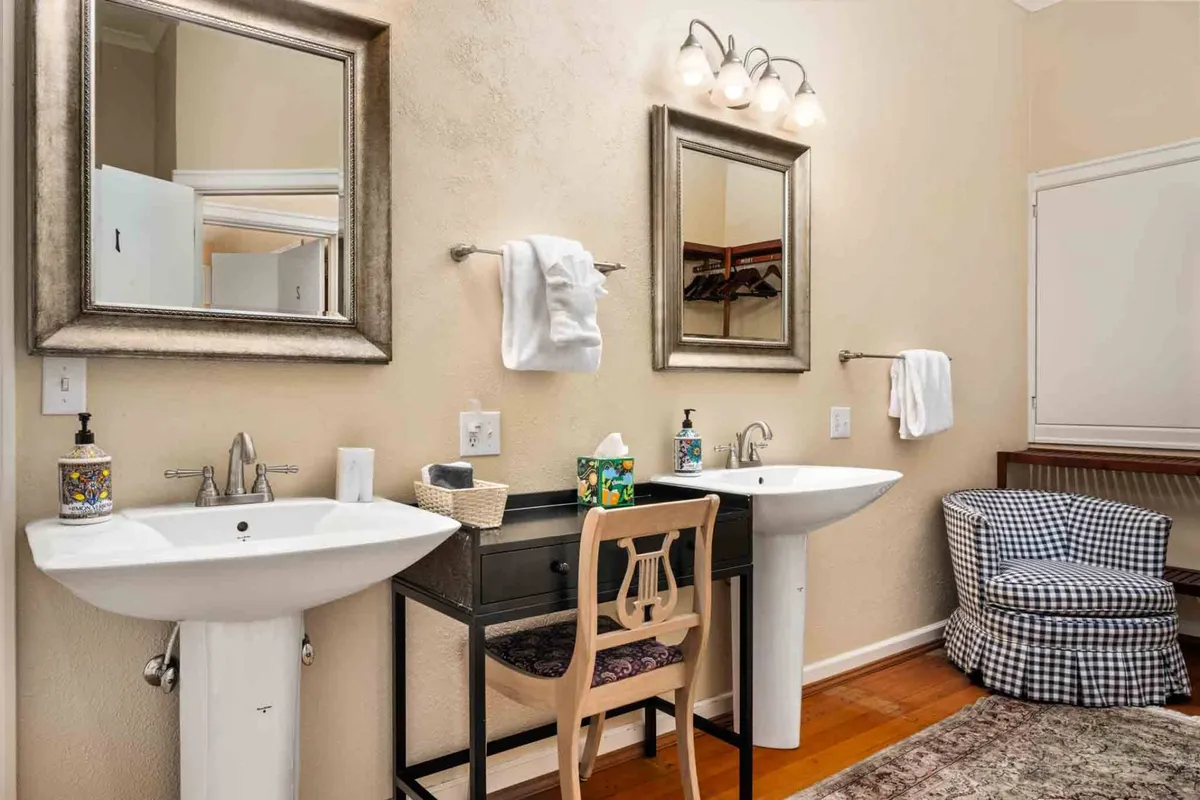 A bright, serene bathroom with double sinks, mirrored cabinets, and comfortable seating. The overall mood is calm and organized, ideal for shared routines while maintaining privacy.
