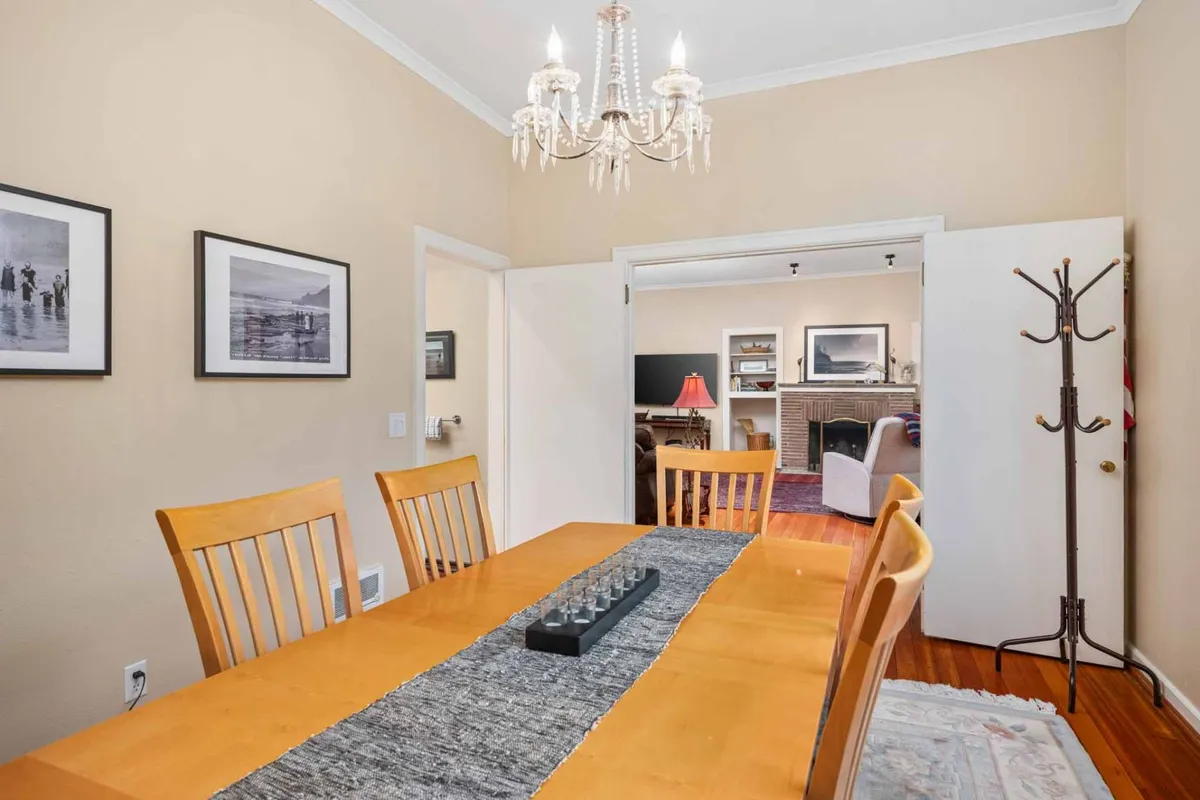 A warm dining area anchored by a large table, elegant chandelier, and tasteful wall art. The space connects visually to a living room beyond, suggesting openness and flow for large groups to dine and chat comfortably.