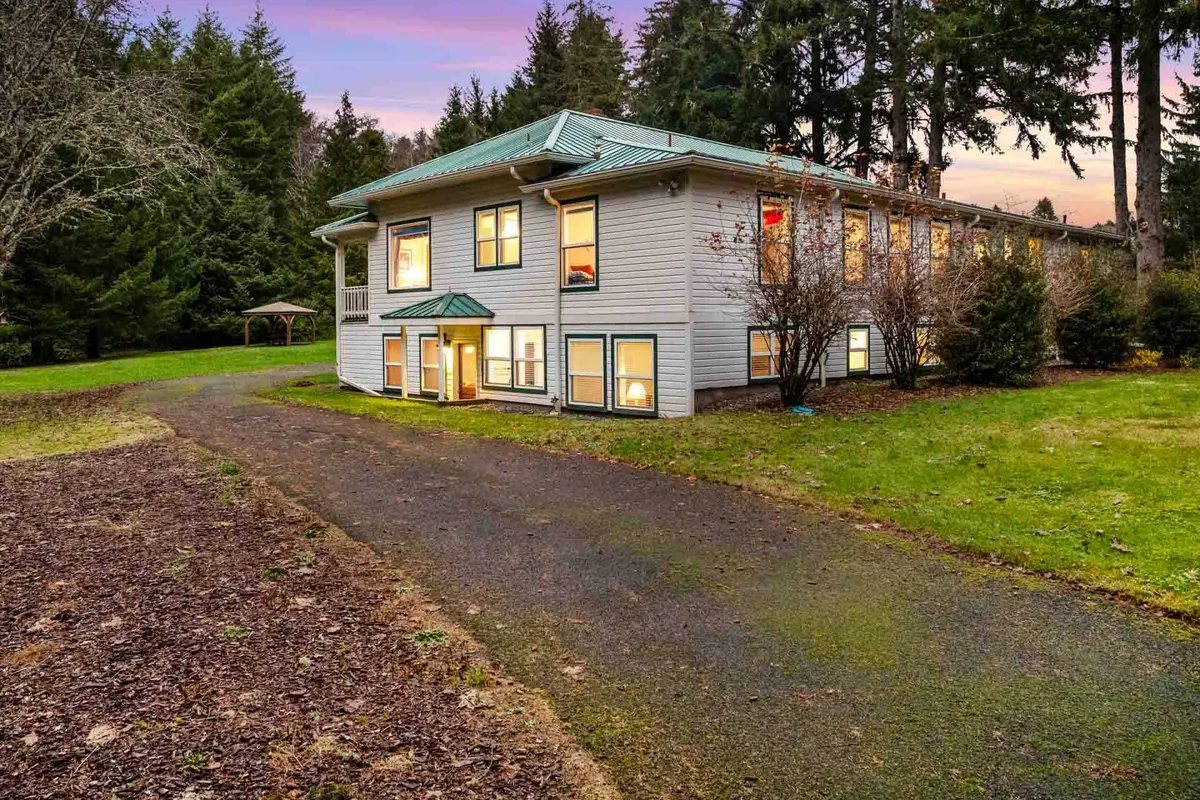 A welcoming exterior view of the Schoolhouse, framed by mature trees and set back from the driveway. The house’s classic facade and peaceful natural surroundings evoke a sense of historic charm and relaxed small-town living. The wide porch hints at communal gatherings and slow mornings.