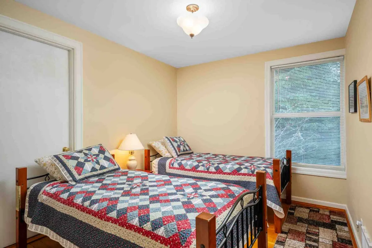 A warm, inviting bedroom scene with two beds covered in quilts, a lamp on a shared nightstand, and a window with blinds letting in natural light. Framed pictures add a homey touch, and the scene feels perfect for families or friends sharing a space.