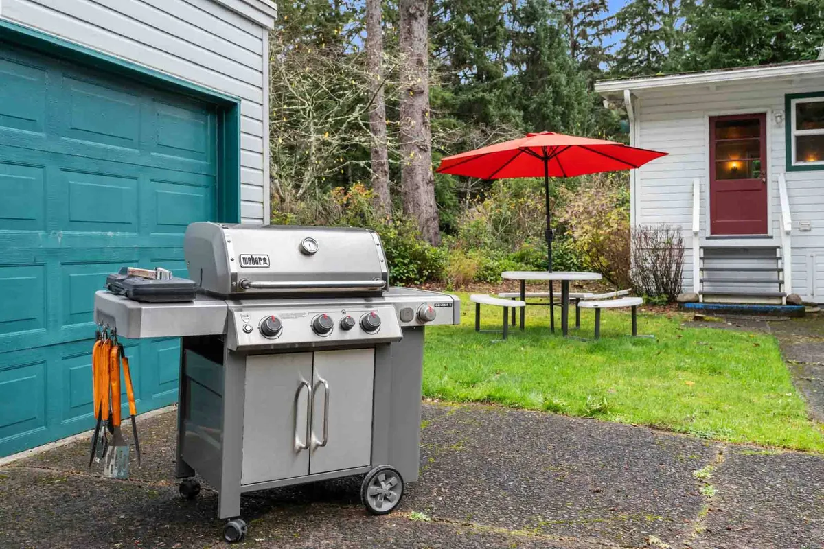 This image captures an outdoor barbecue area with a grill, a picnic table beneath a vibrant red umbrella, and the spaciousness of the surrounding yard. The mood is friendly and communal, signaling the potential for outdoor dining and group activities.